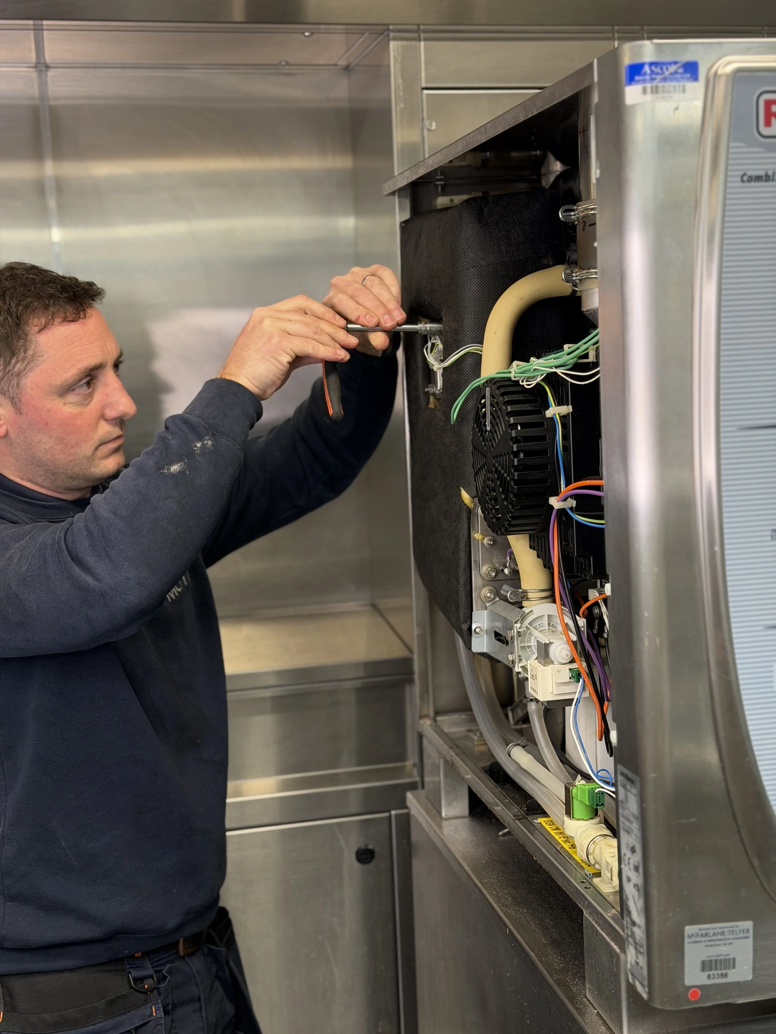 A technician repairing or installing electrical components inside a kitchen appliance, possibly a dishwasher or a washing machine, in a commercial or industrial kitchen setting.