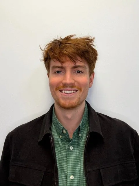 A young man with red hair, beard, and mustache smiling, wearing a green checkered shirt and a black jacket, standing against a plain white wall.