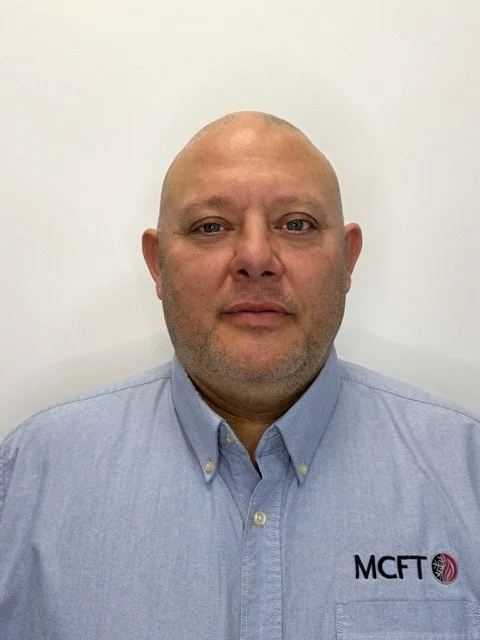 Headshot of a man wearing a light blue collared shirt with an MCFT logo, standing against a white background.