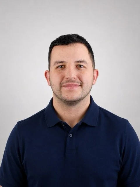 Portrait of a young man with short dark hair, light skin, wearing a navy blue collared shirt, smiling softly against a plain light gray background.