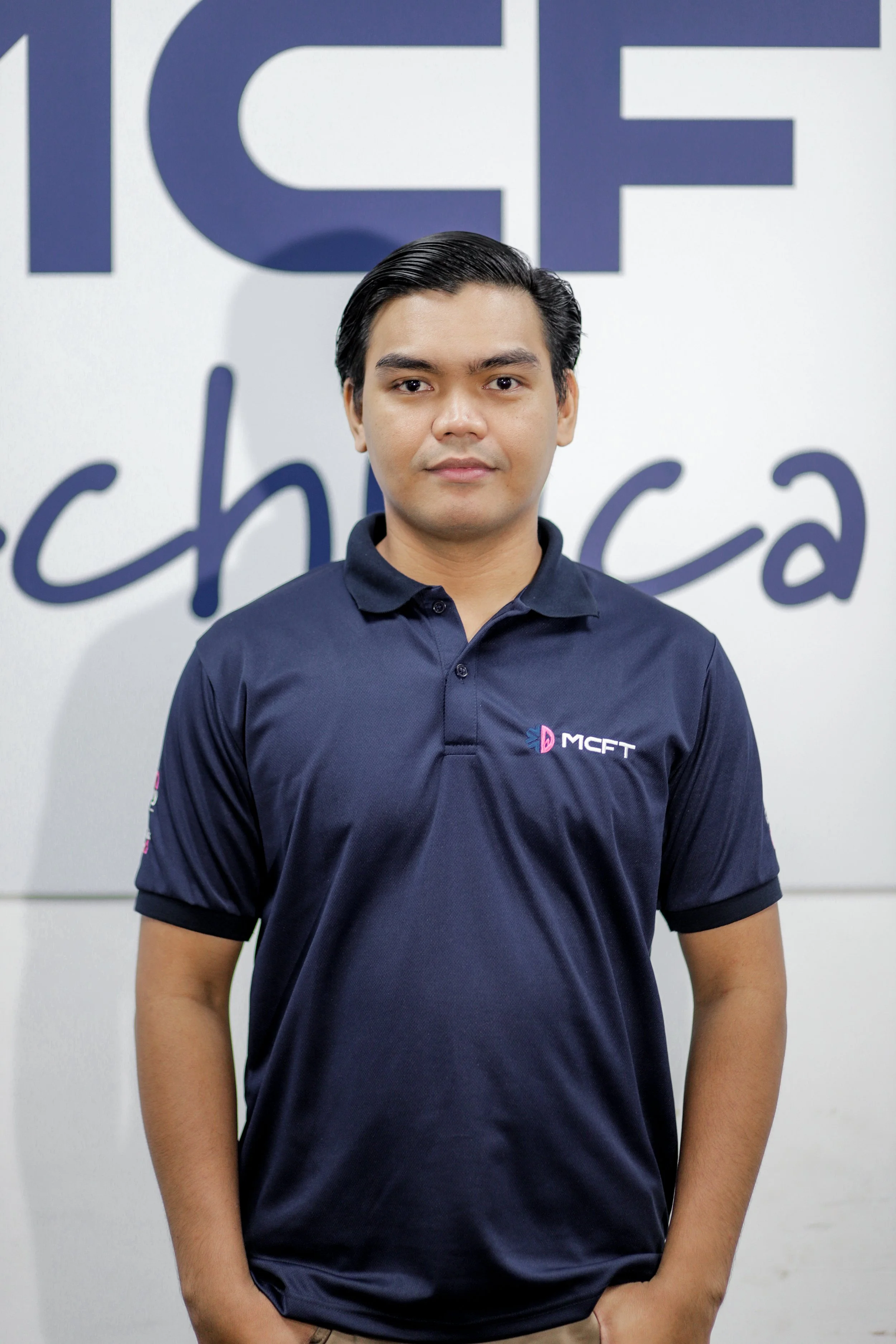 A young man with black hair in a formal business casual outfit standing against a wall with a logo and text background.