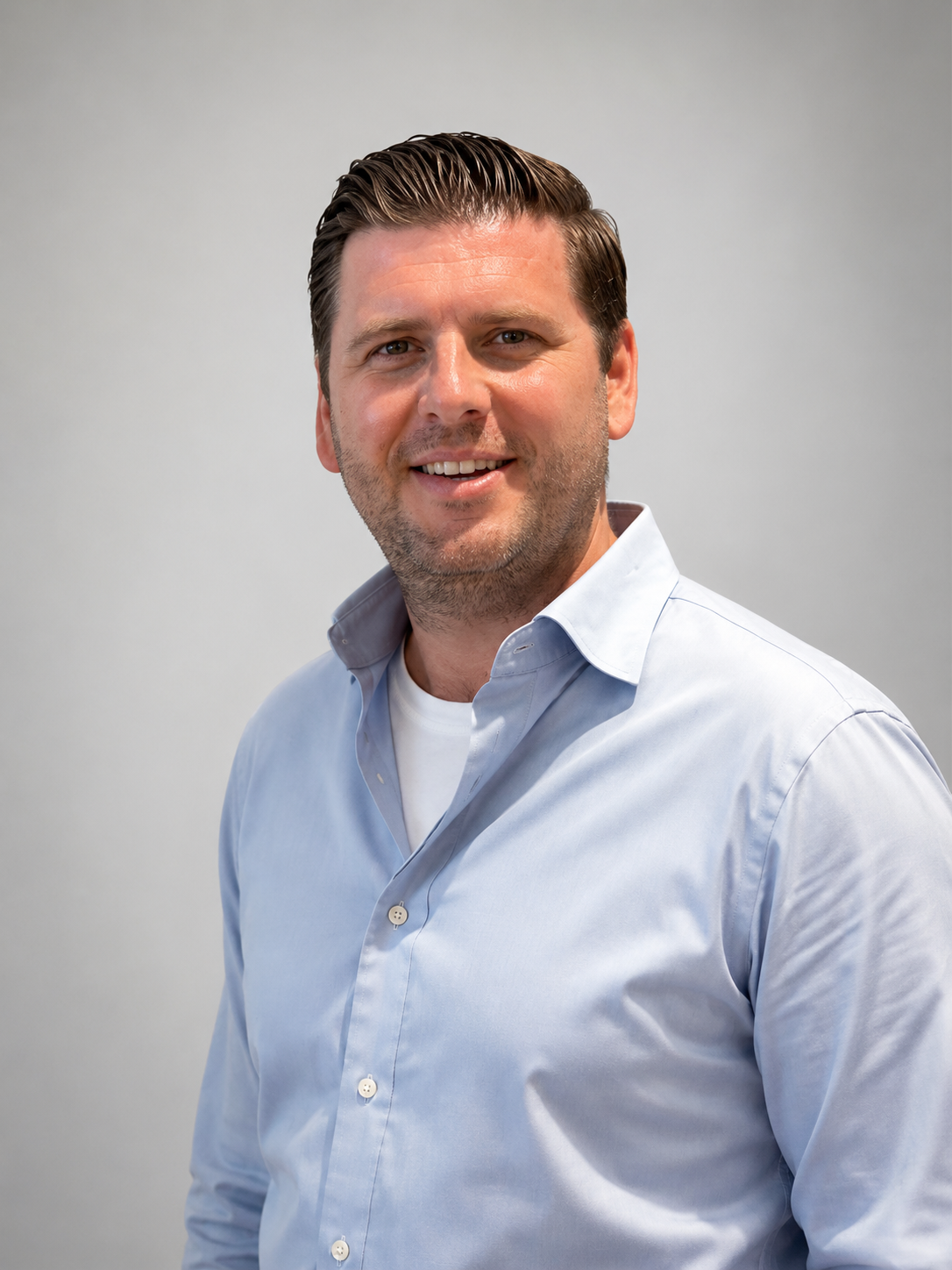 Headshot of a smiling man with light brown hair, wearing a light blue button-up shirt over a white t-shirt, standing against a plain light gray background.
