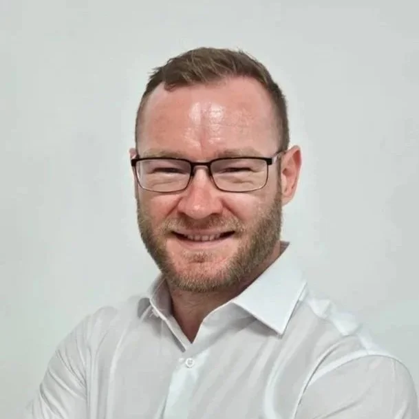 A man with glasses, short brown hair, and a beard smiling and wearing a white collared shirt against a plain white background.