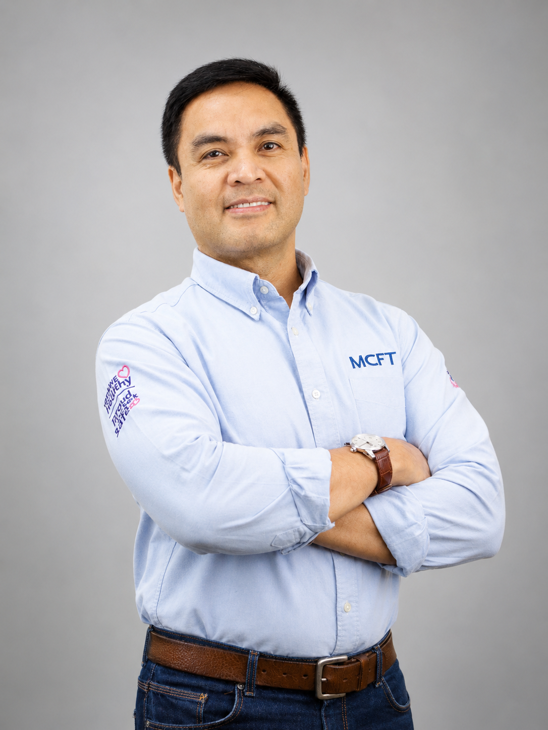 A man with dark hair and light brown skin wearing a light blue button-up shirt with patches and white embroidered lettering, crossing his arms, against a plain light gray background.