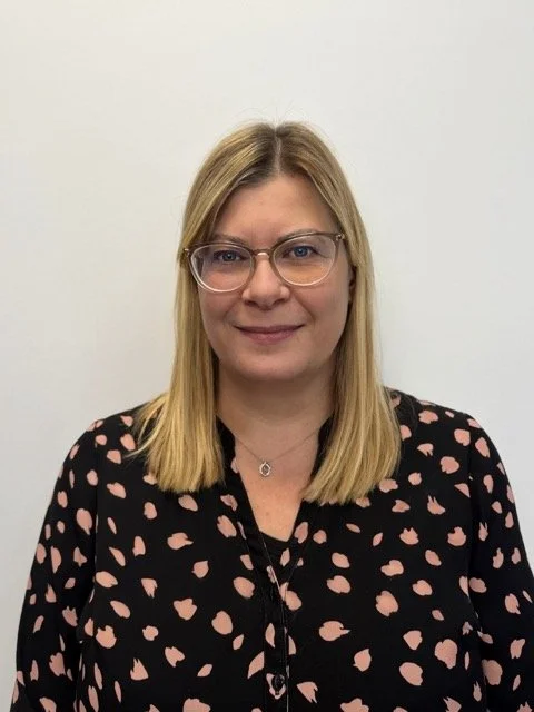Portrait of a woman with shoulder-length blonde hair, wearing glasses, a black blouse with pink spots, and a necklaces, standing against a plain white wall.