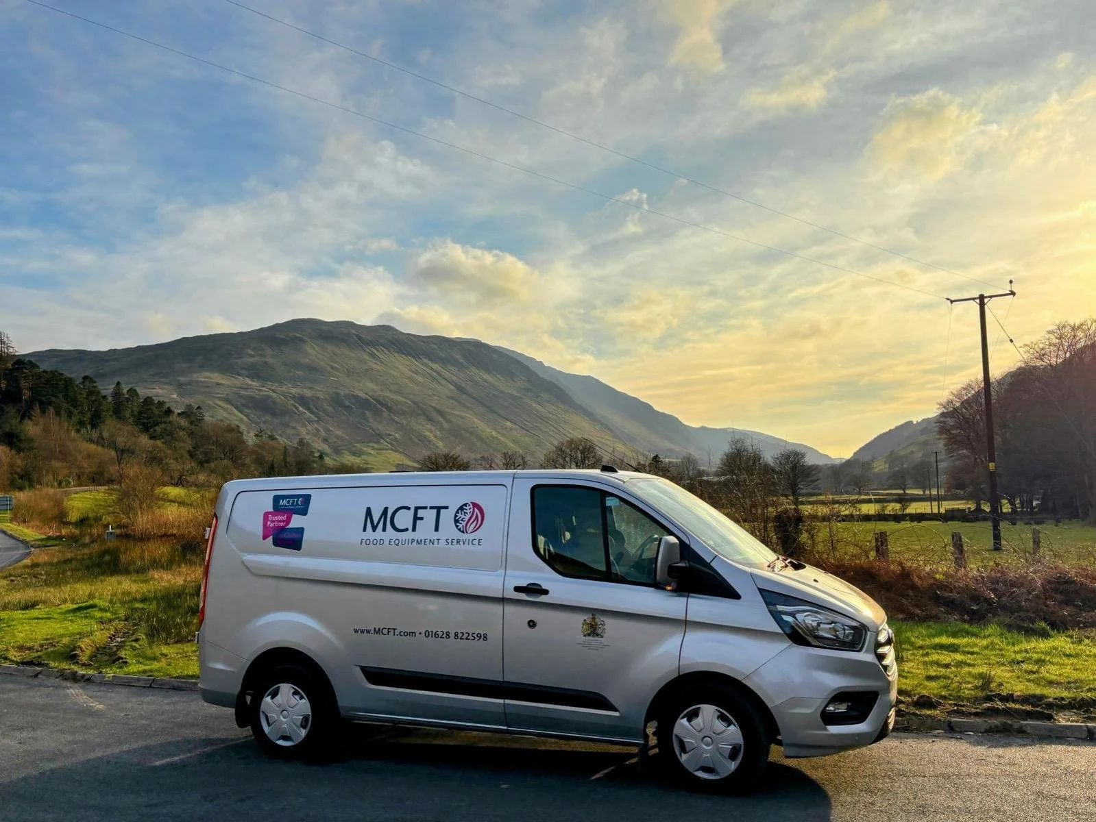 A white van with MCFT Food Equipment Service logo parked on a rural road with green grass, trees, telephone poles, and mountainous landscape in the background during sunset or sunrise.
