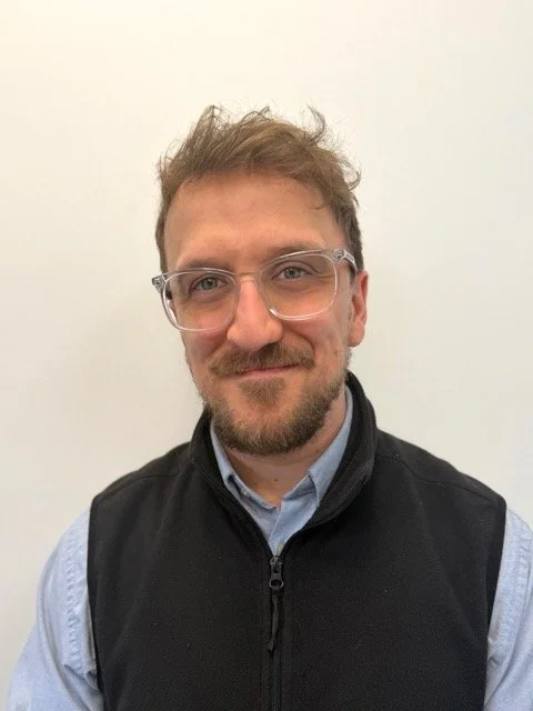 A young man with glasses, a beard, and curly hair smiling in front of a plain white wall, wearing a black zip-up vest over a light blue collared shirt.