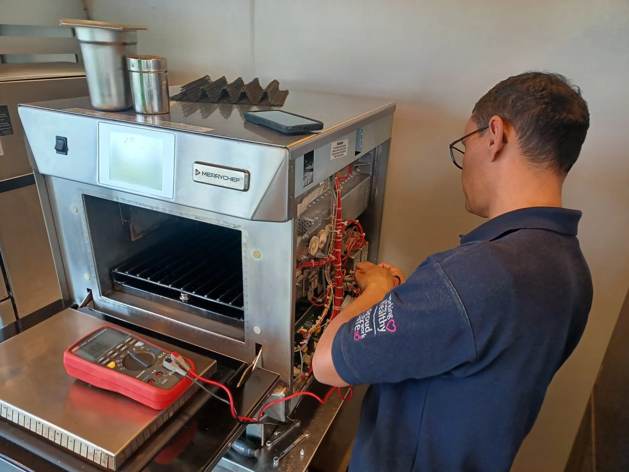 A man working on a commercial oven or kitchen equipment, with exposed wiring inside. There are tools and a digital multimeter placed on the counter in front of him.