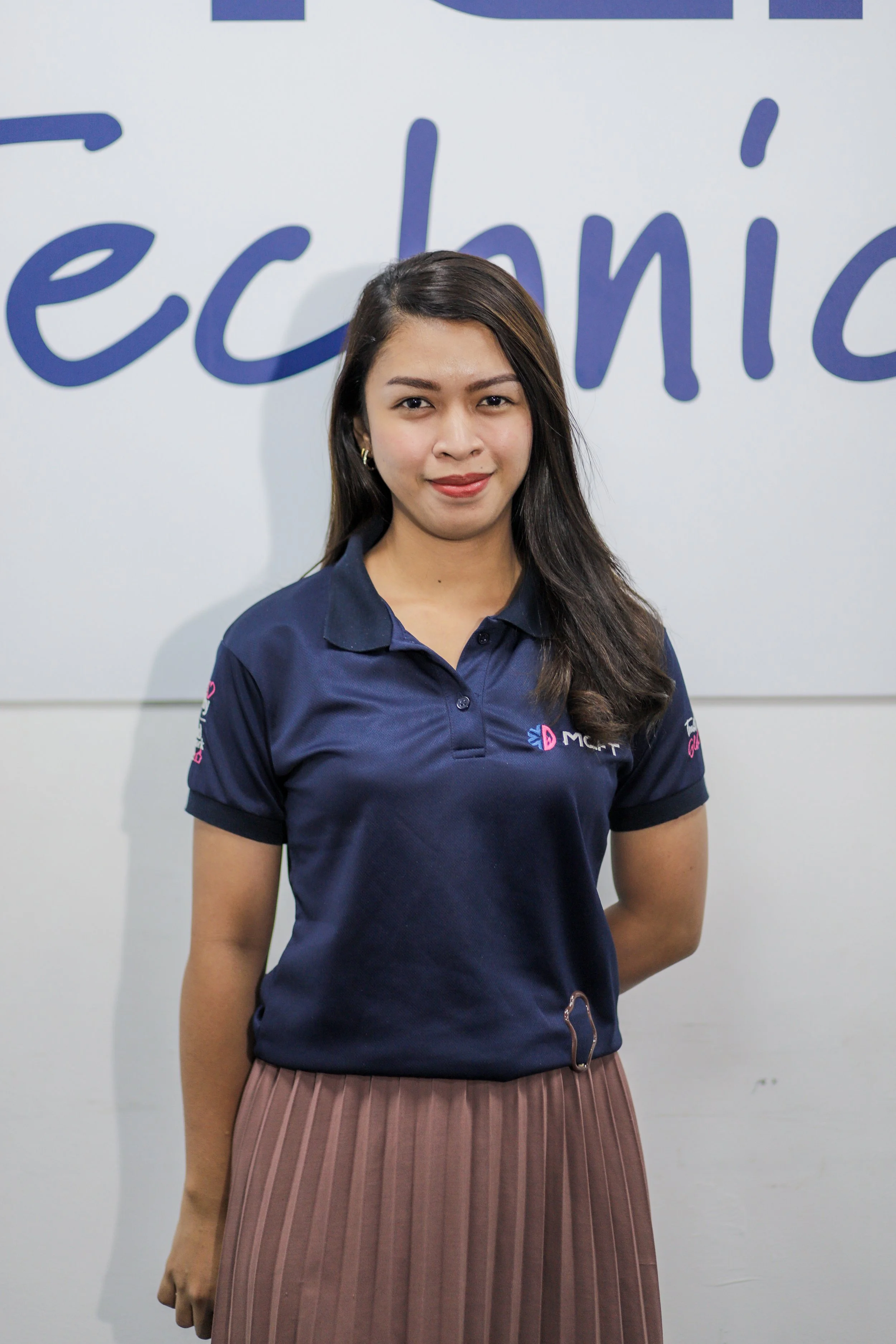 A young woman with long dark hair, wearing a navy blue polo shirt with logos and a pleated skirt, standing in front of a sign that says 'ec.'