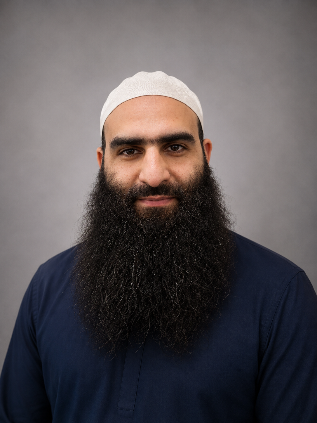 A man with a dark beard wearing a white prayer cap and a navy blue shirt, standing against a gray background.