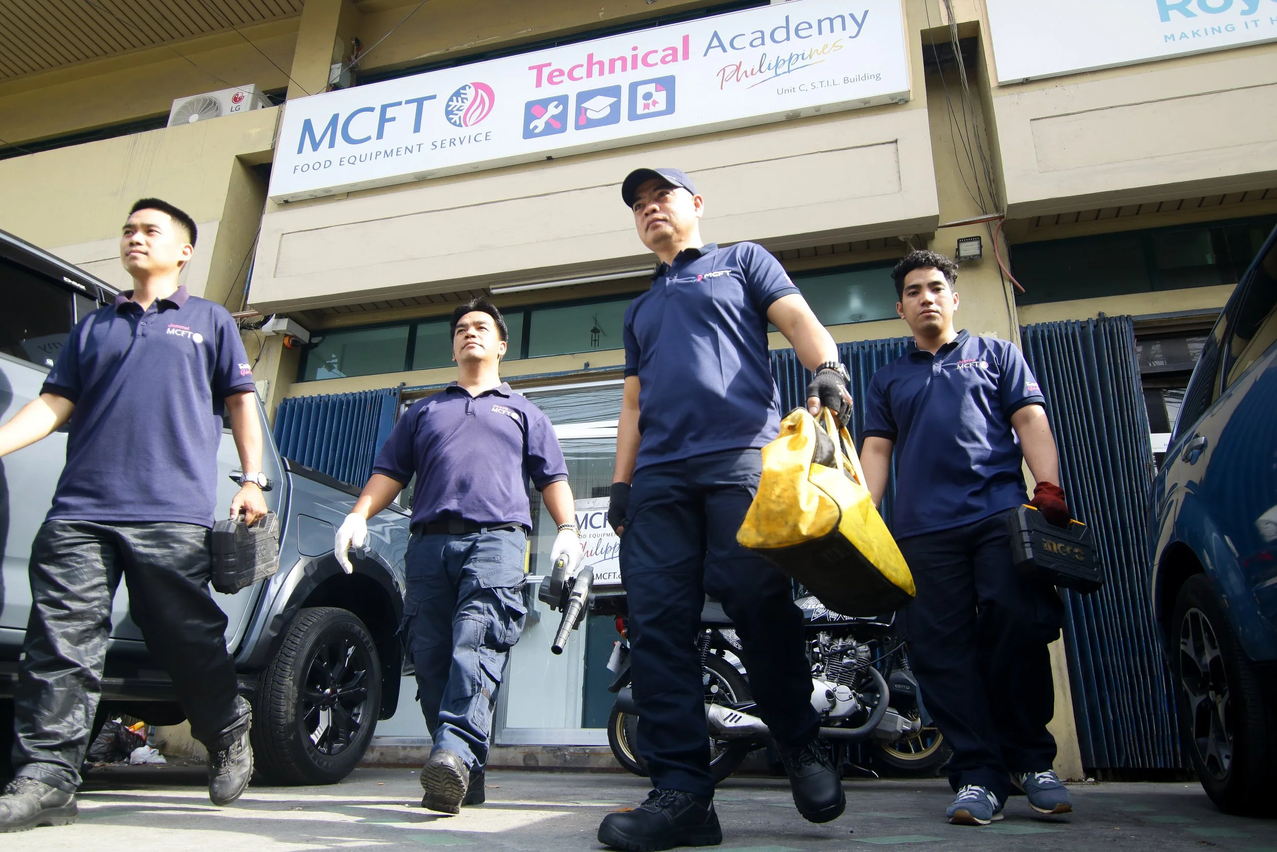 Four workers wearing blue uniforms with 'MCFT' logo walking in front of a building with signs for 'MCFT Food Equipment Service' and 'Technical Academy Philippines'. Some workers carry tools and equipment.