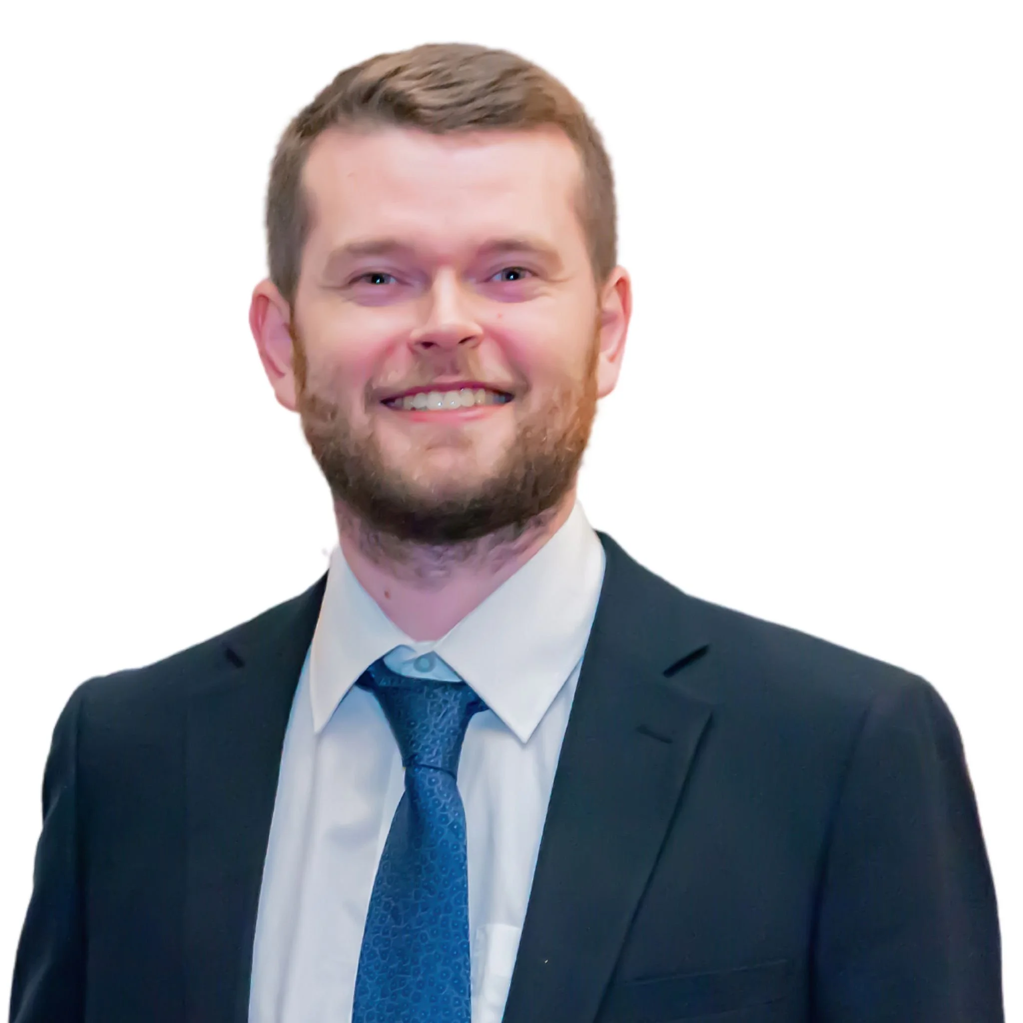 Close-up of a smiling young man with light brown hair, beard, dressed in a black suit, white shirt, and blue tie, standing against a white background.