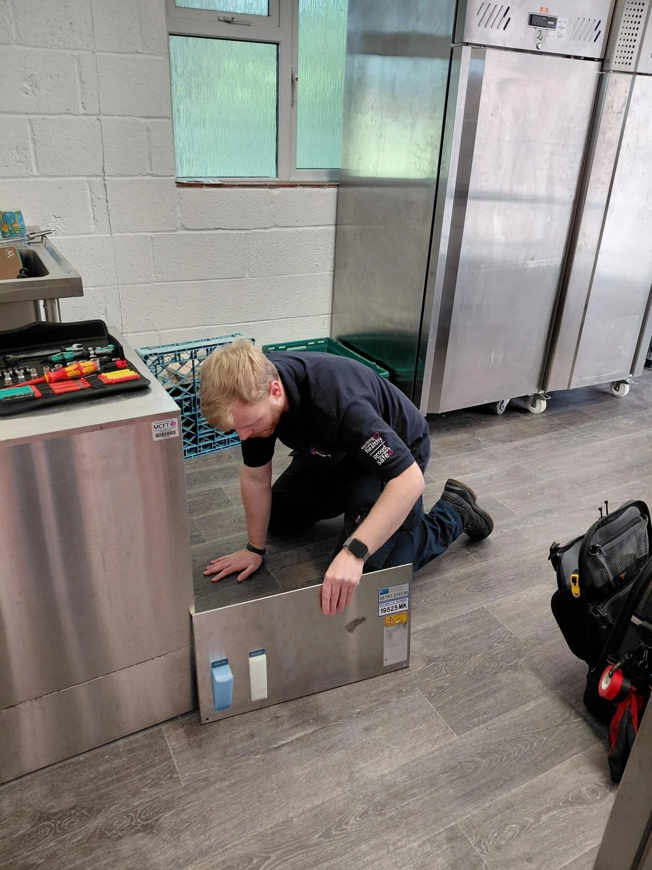 A man kneeling on the floor working on a large stainless steel appliance in a kitchen or utility room, with tools and equipment nearby.