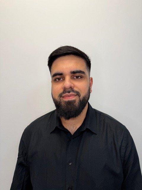 A young man with a beard and short dark hair, wearing a black button-up shirt, standing against a plain white wall.
