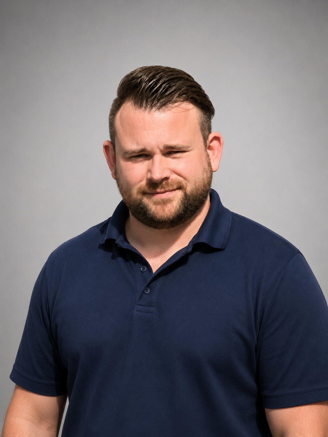 A man with a beard and short dark hair wearing a navy blue collared shirt against a plain gray background.