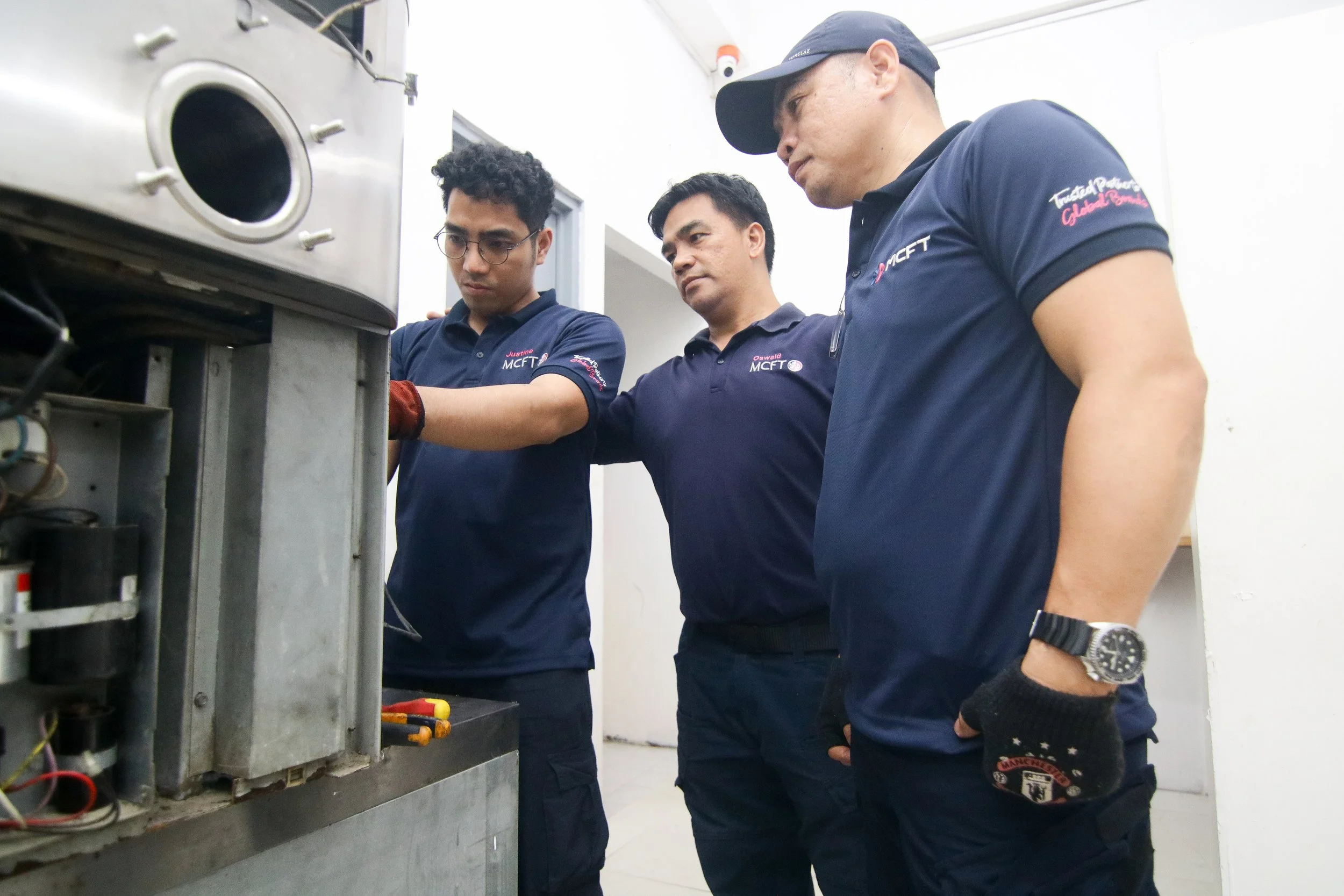 Three men in navy blue shirts working on a machine in a white-walled room.