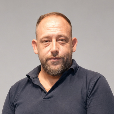 A man with a beard and short hair wearing a dark polo shirt, standing against a plain, light-colored background.