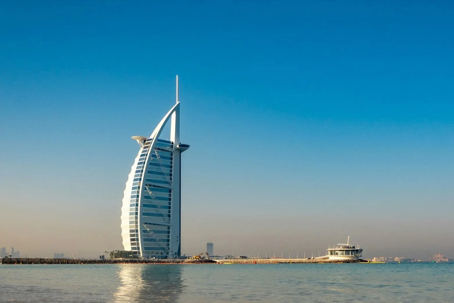 View of the Burj Al Arab hotel in Dubai with water in the foreground and a clear blue sky.