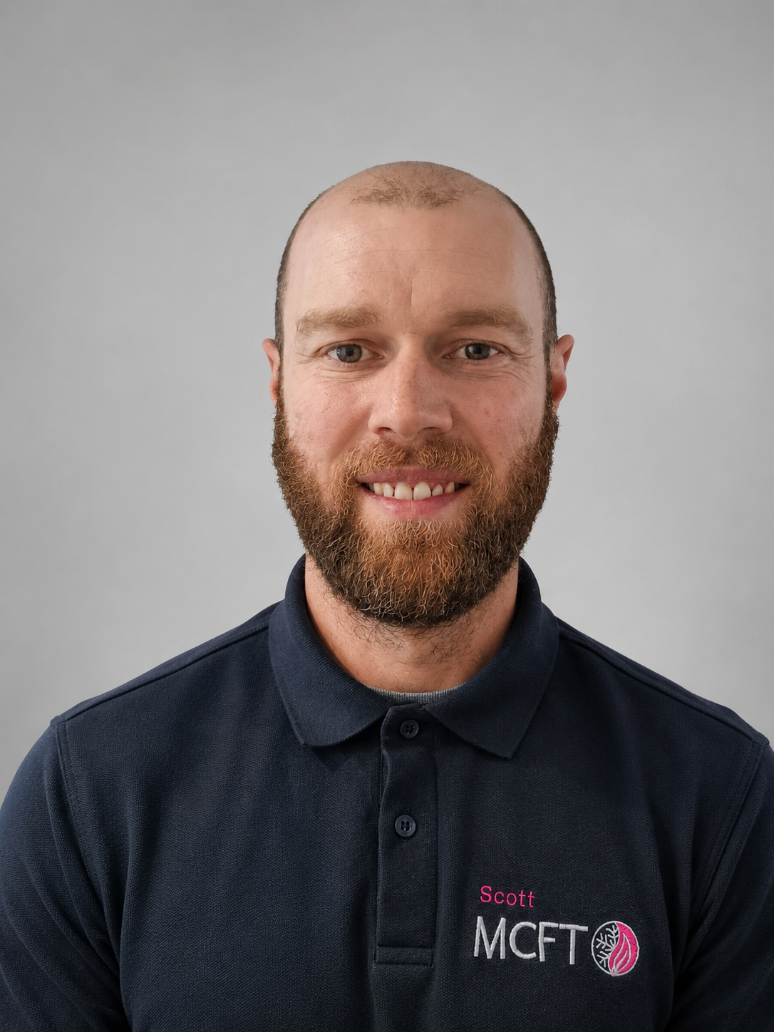 A man with a beard and short hair, smiling, wearing a navy blue polo shirt with a company logo that reads "SCOTT MCFT" on a plain gray background.
