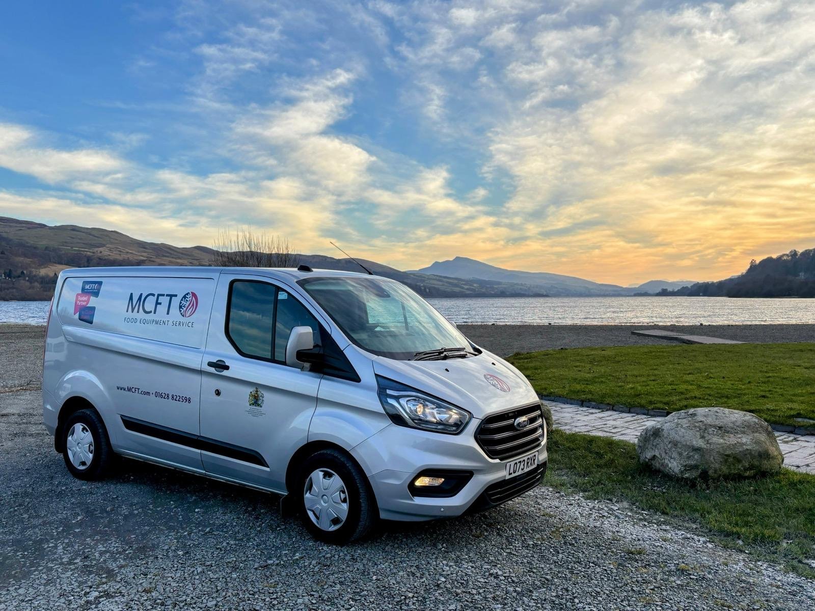 A white van for MCFT Food Equipment Service parked by a lake at sunset, with mountains and a cloudy sky in the background.