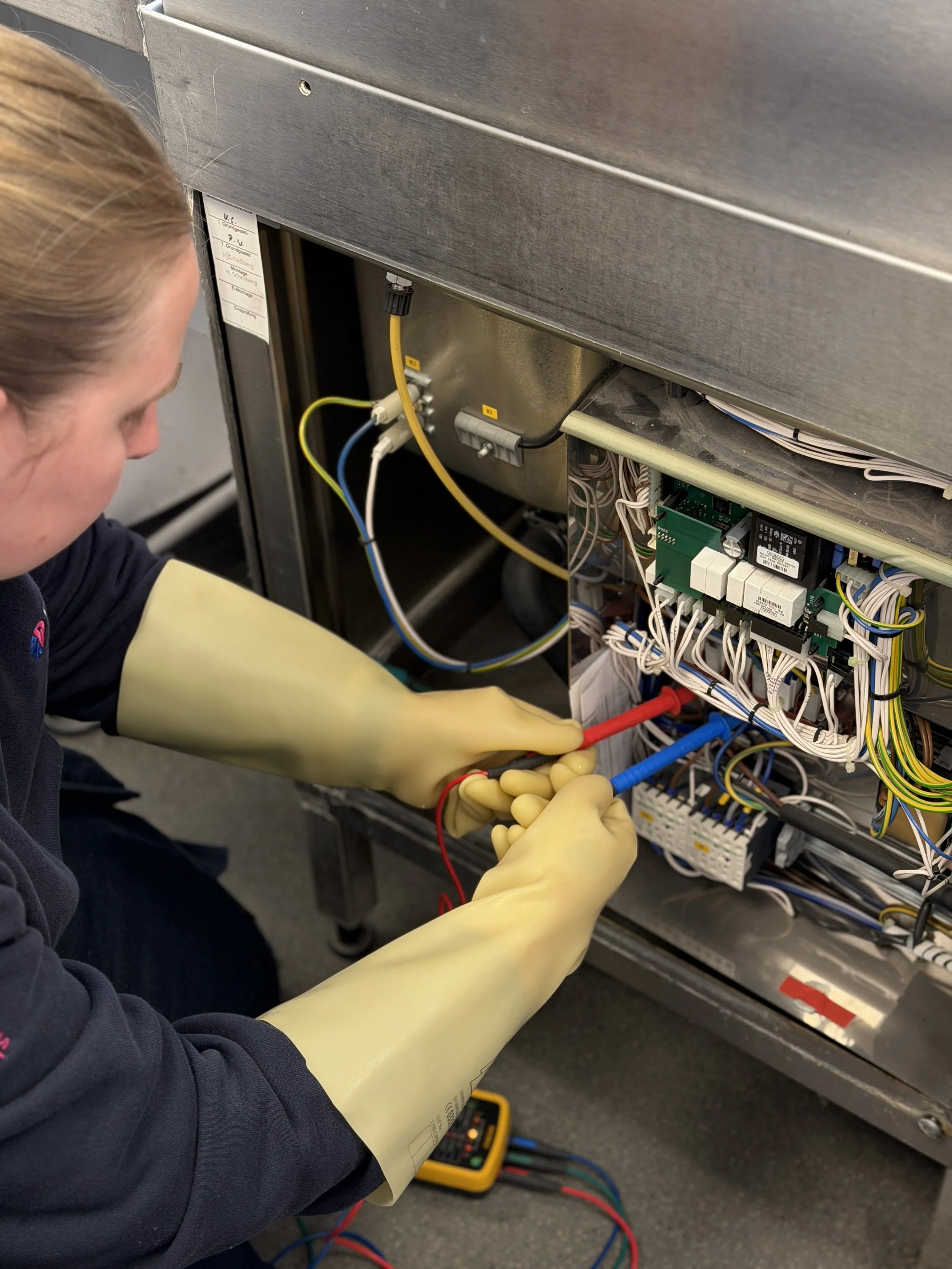 A technician wearing yellow rubber gloves is working on the internal components of an electrical or electronic unit, using tools, with a multimeter and colorful wires visible nearby.