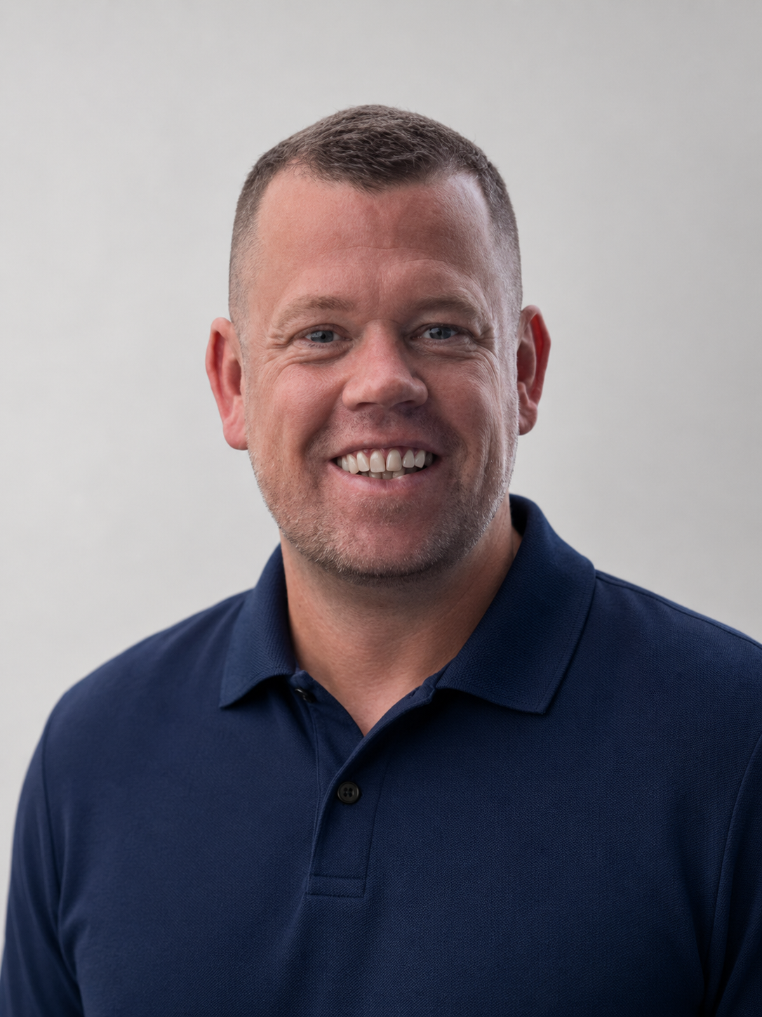 A smiling man with short brown hair wearing a navy blue polo shirt against a plain light gray background.
