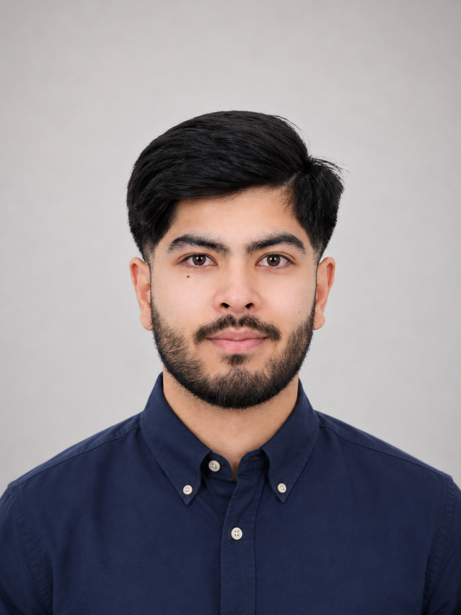 Portrait of a young man with dark hair and a beard wearing a dark blue button-up shirt, looking at the camera with a neutral expression, against a plain light gray background.