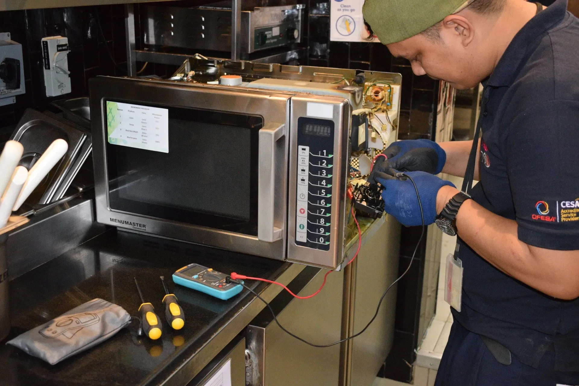 A worker in a dark navy uniform with a green beanie and gloves is repairing a microwave oven. The microwave's front panel is open, exposing the internal components and wiring. On the countertop, there are screwdrivers, an energy meter, and a cloth. In the background, there are kitchen utensils and a sign with cleaning instructions.