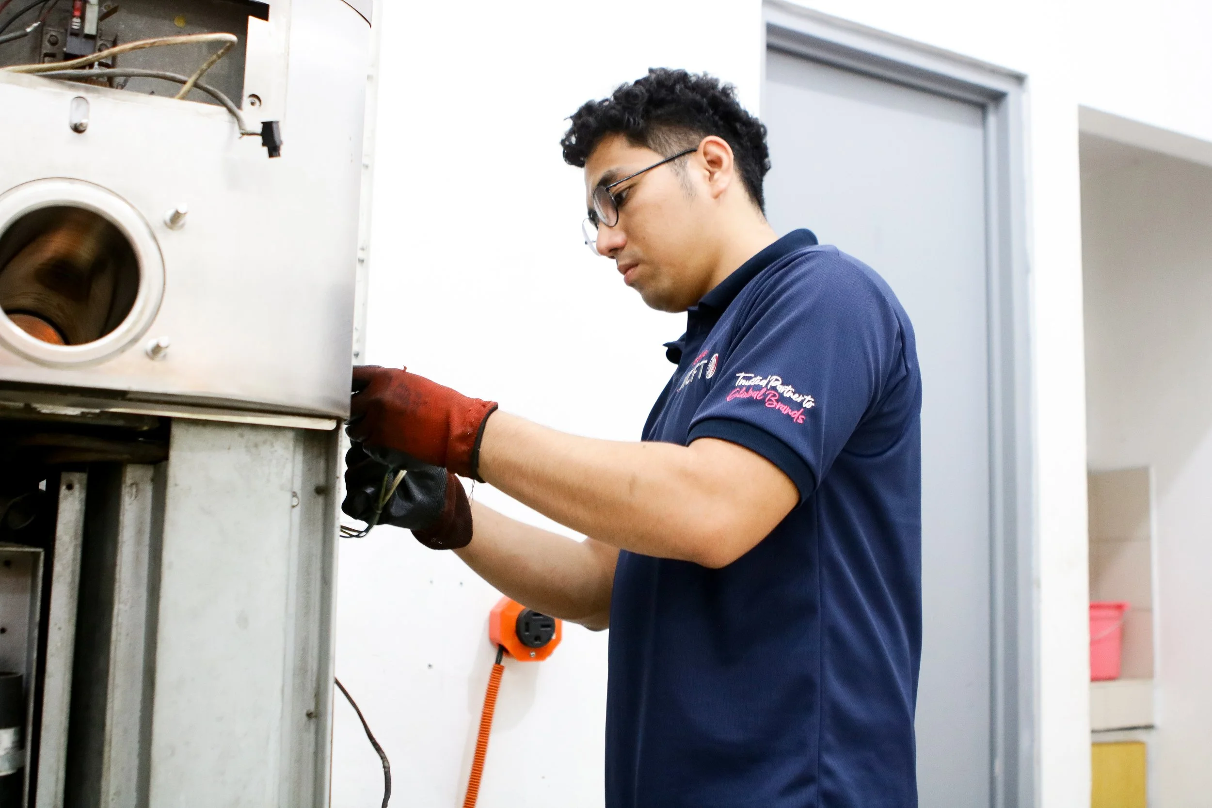 A technician working on electrical/air conditioning equipment in an indoor setting, wearing safety gloves and a navy blue uniform.