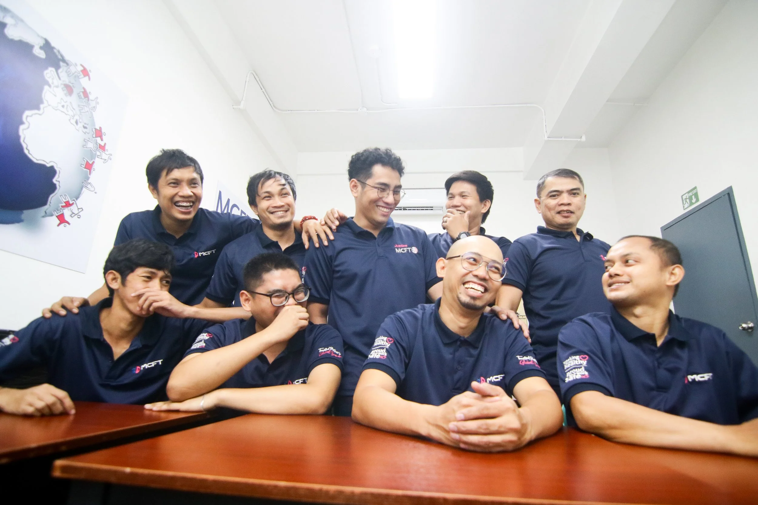 Group of men in dark blue polo shirts sitting and standing around a wooden table in an office or conference room, smiling and enjoying each other's company.