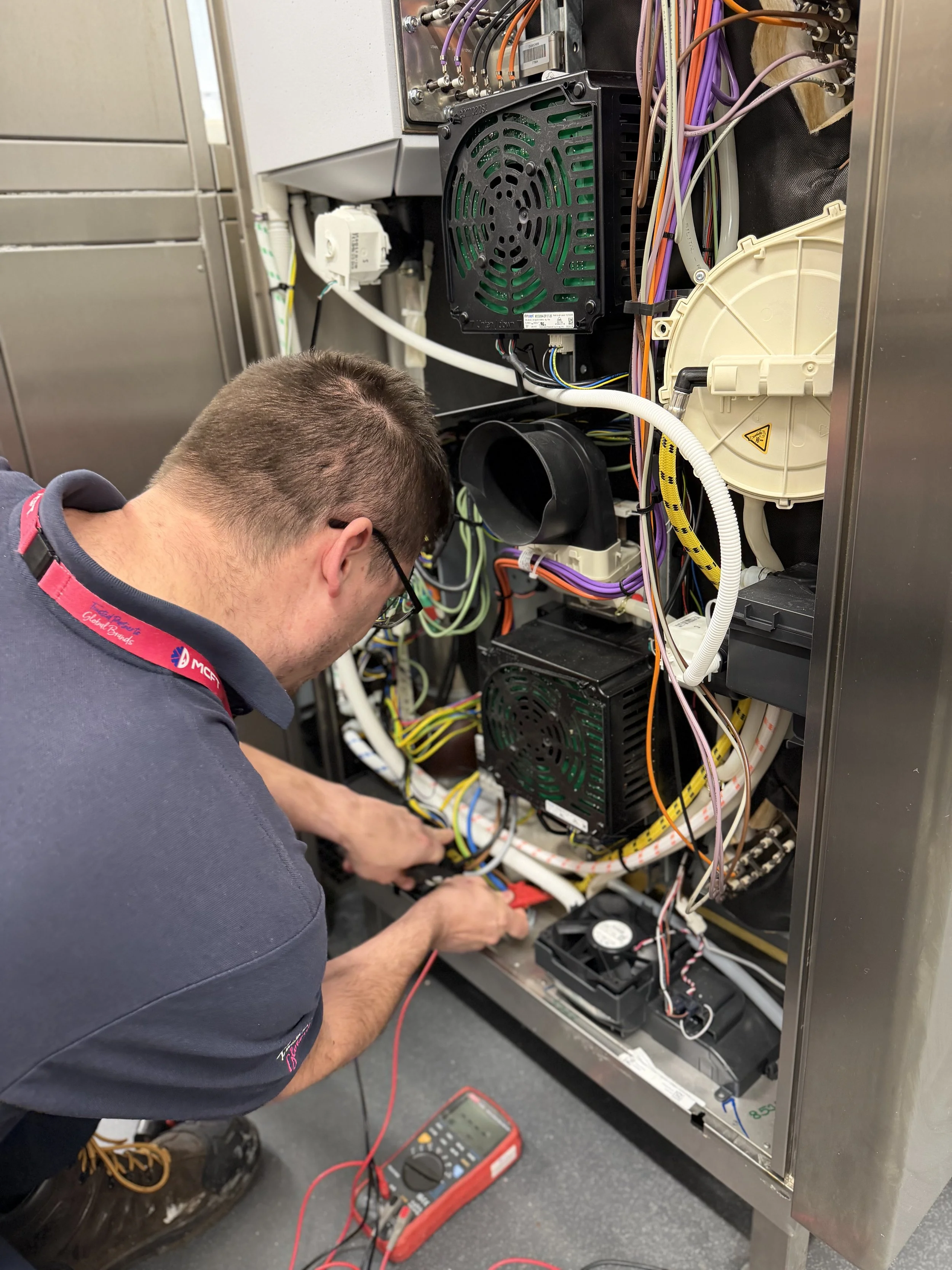 Technician working on an industrial electrical panel with wires and components inside a metal cabinet, using a multimeter for testing.