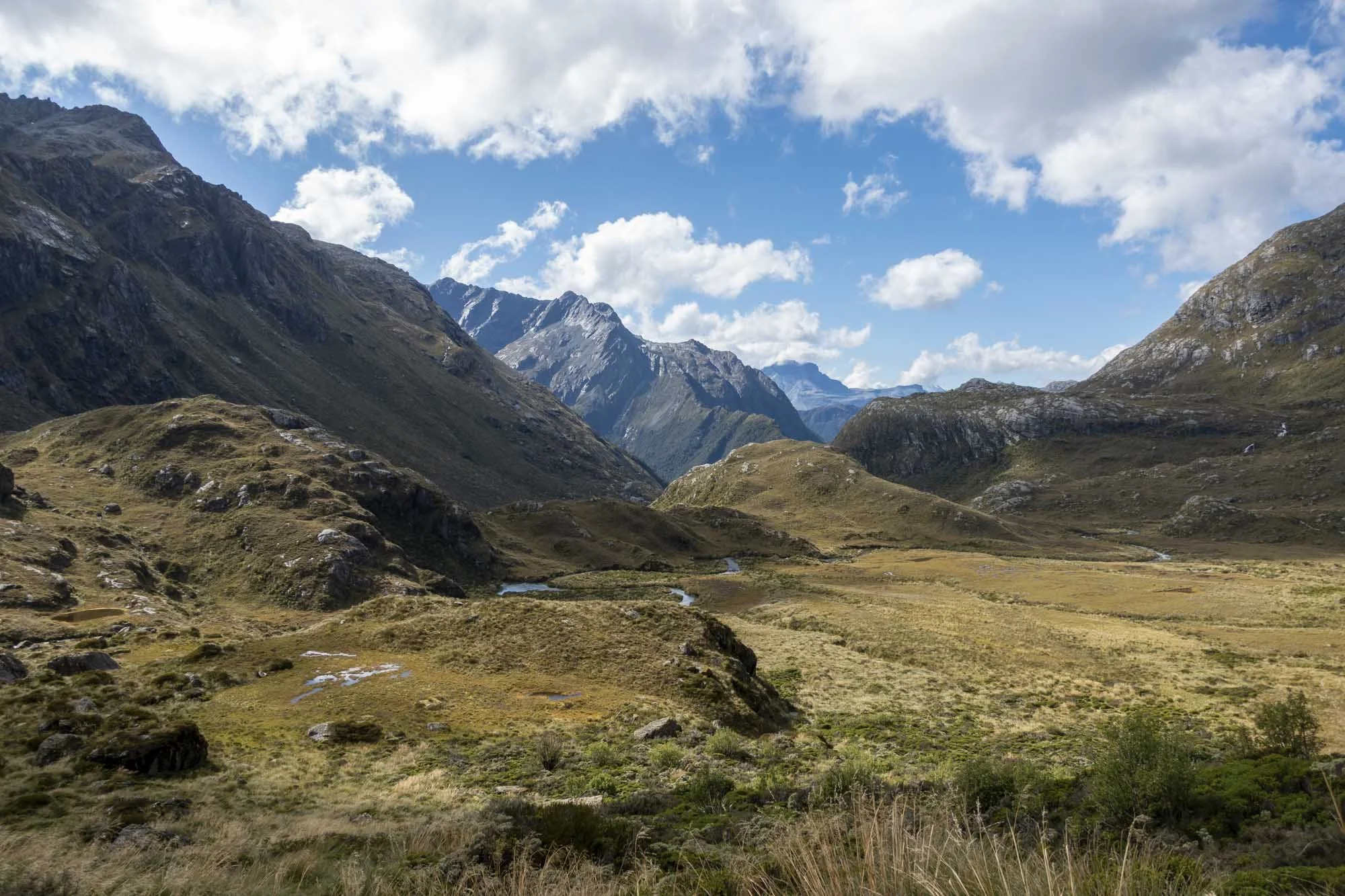 Scenic view of a mountain valley with grassy hills, rocky slopes, and patches of small lakes, under a partly cloudy sky.