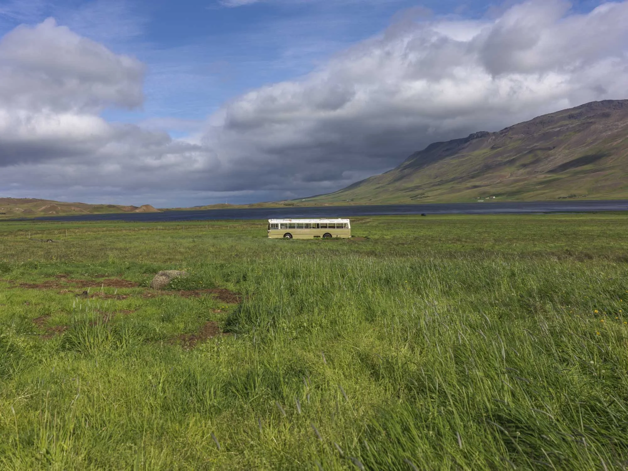 A bus parked in a vast green field with mountains and a lake in the background under a partly cloudy sky.