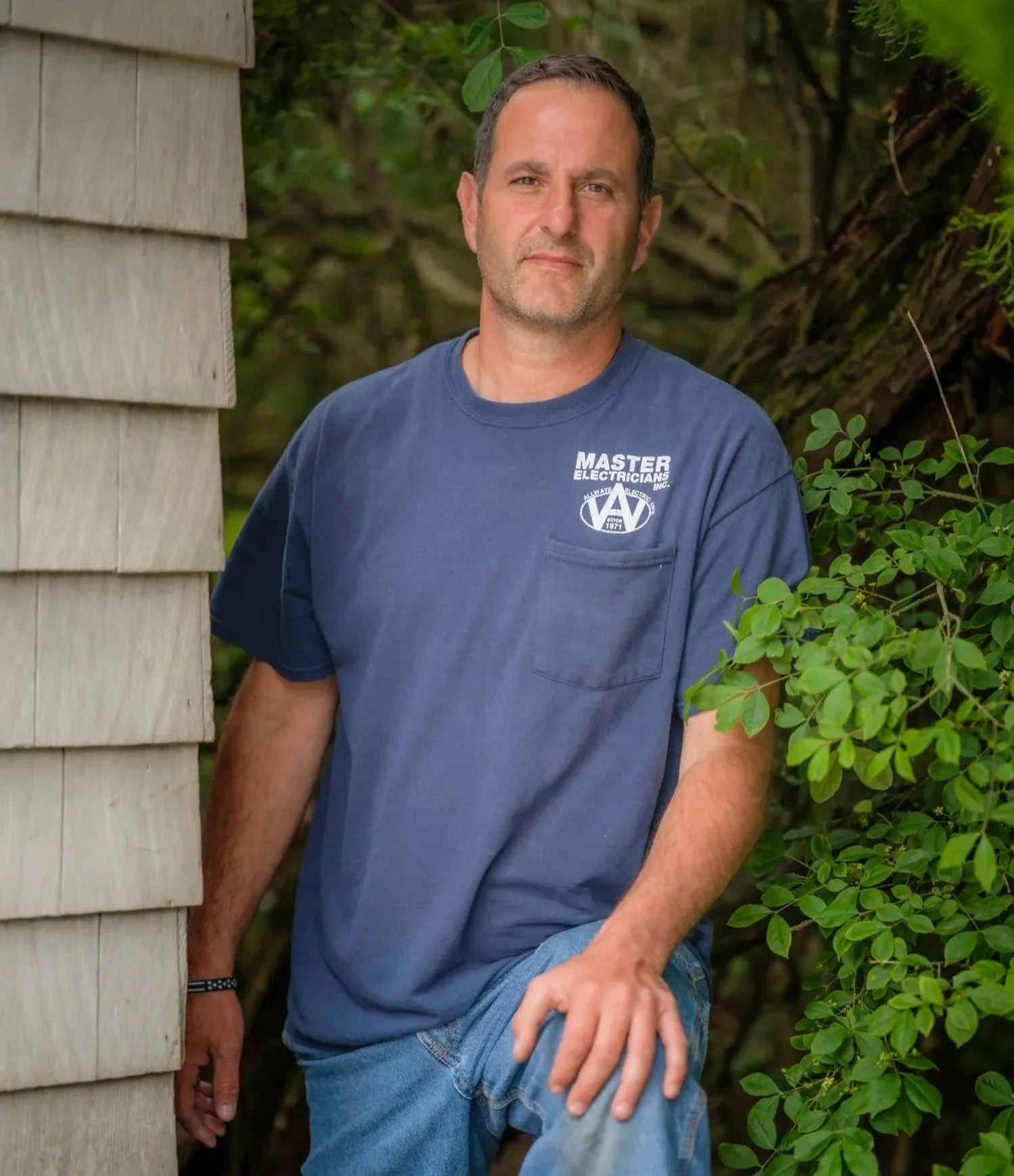 A man in a blue t-shirt with a logo reading 'Master Electricians' stands outdoors, leaning against a wooden structure with green foliage around him.
