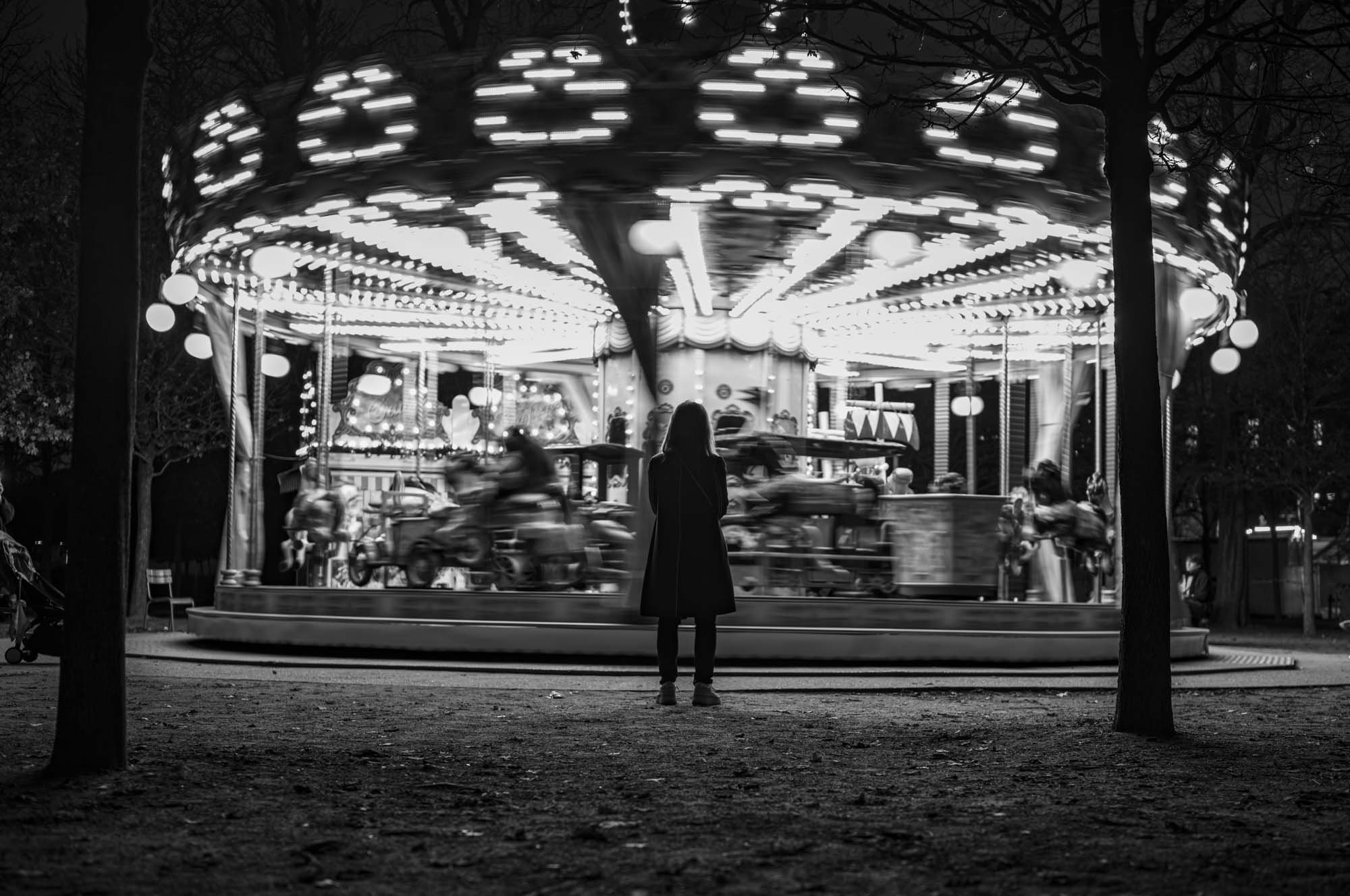 A person standing in front of a moving carousel ride at night, with blurred motion of the ride's lights and figures.