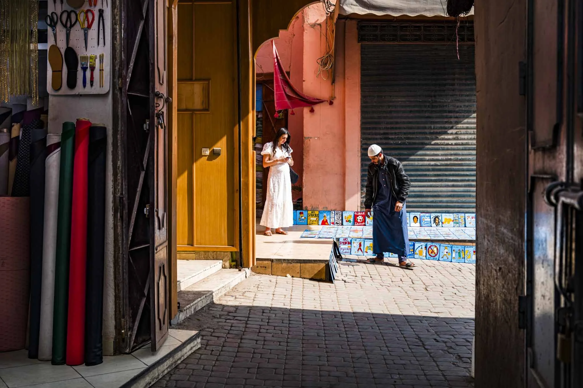 View through an open shop door onto a street scene with a woman in a white dress looking at a phone and a man in traditional attire looking at items displayed outside.