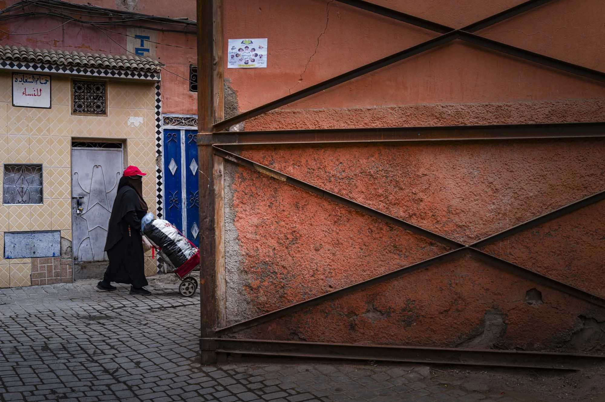 A woman wearing a black cloak, black face mask, and red cap is pushing a pushcart with bags on it on a cobblestone street. Behind her is a beige tiled building with a small window, a door with a star design, and a sign in Arabic. The street is enclos
