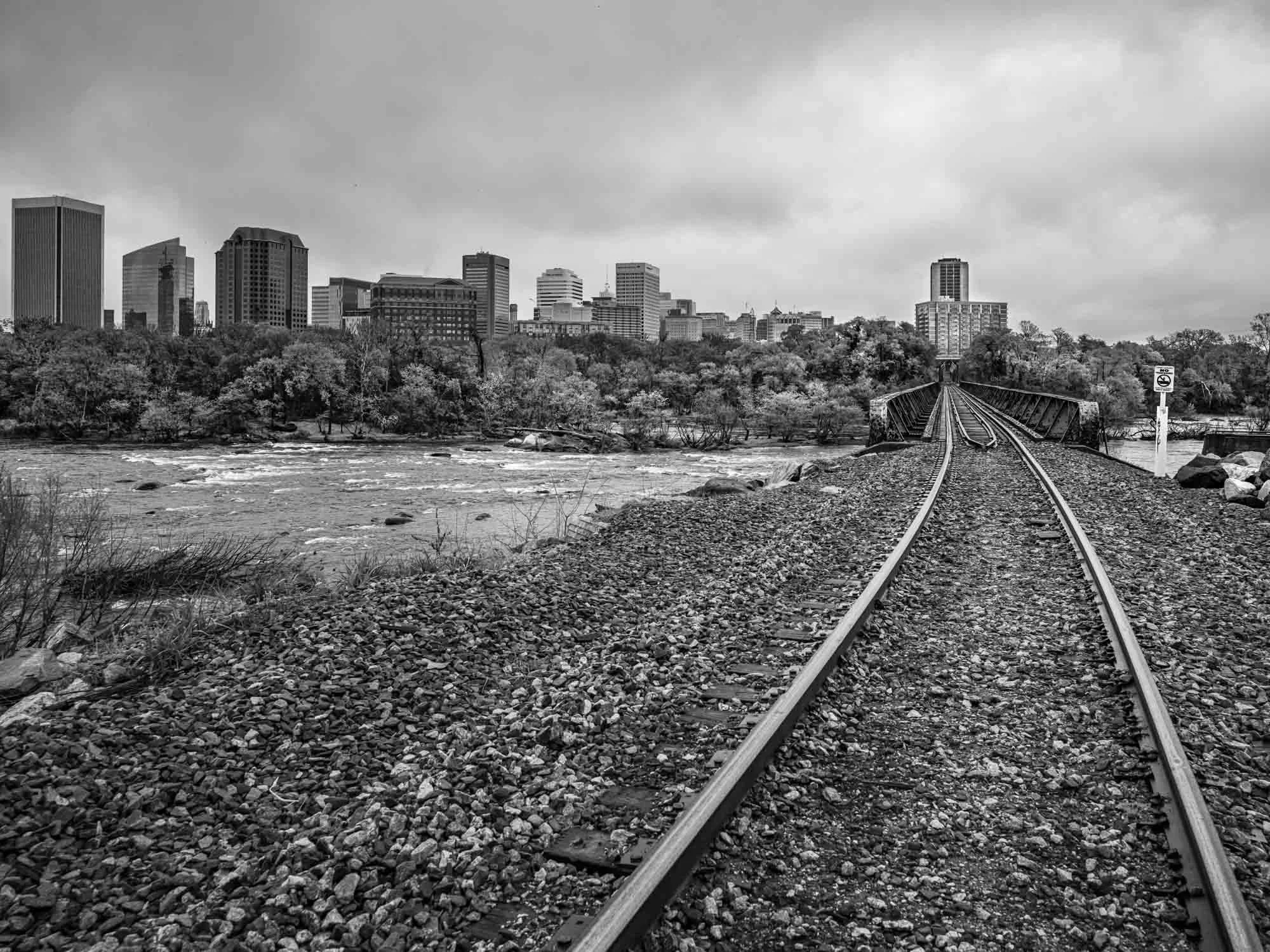 Black and white photo of a railway track leading to a city skyline with tall buildings, trees, and a river in the foreground.