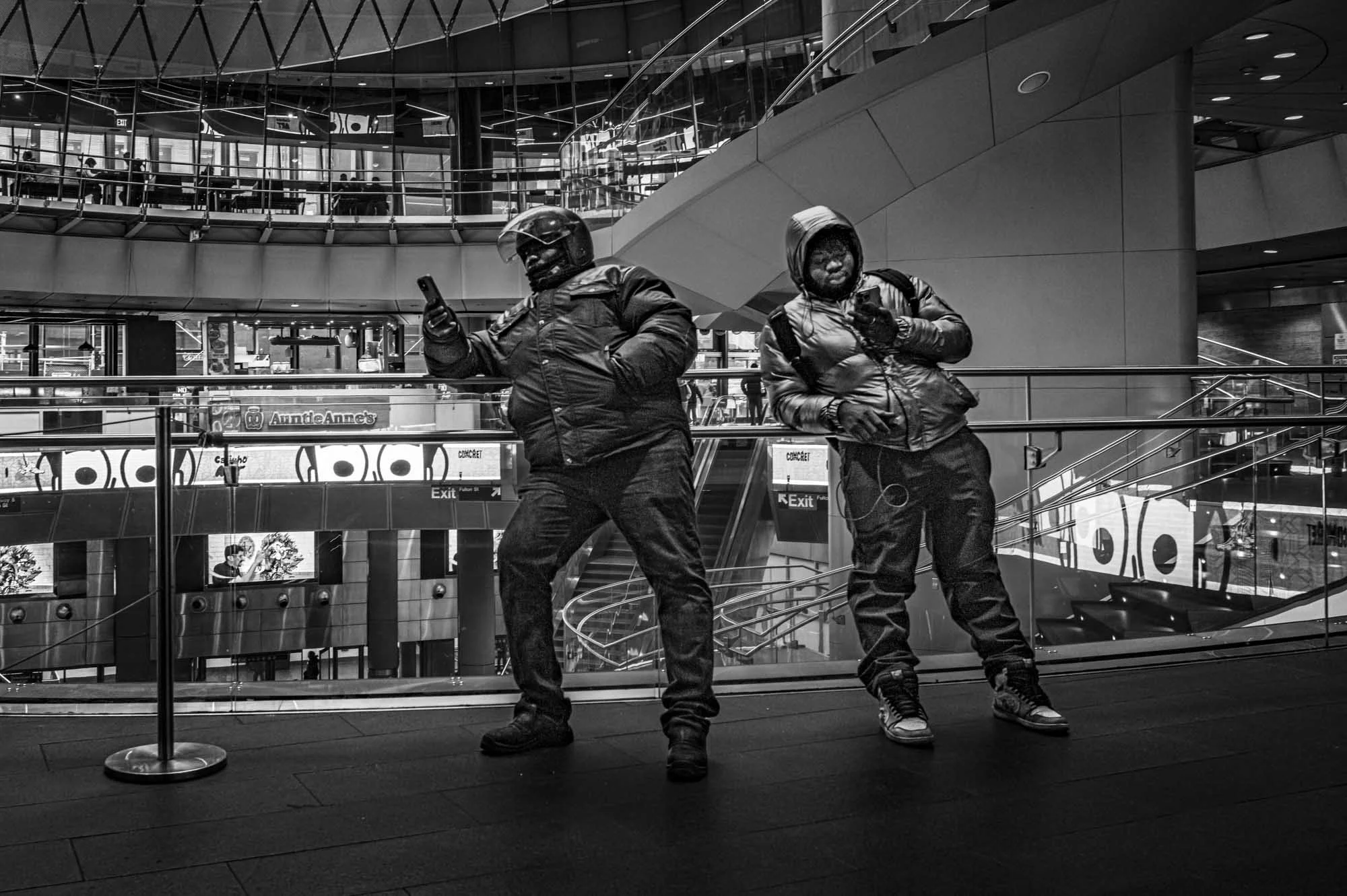 Two individuals in protective jackets, pants, and gloves standing inside a modern, multi-level shopping mall, one using a smartphone and the other looking at a device, with glass railings, escalators, and store signs in the background.