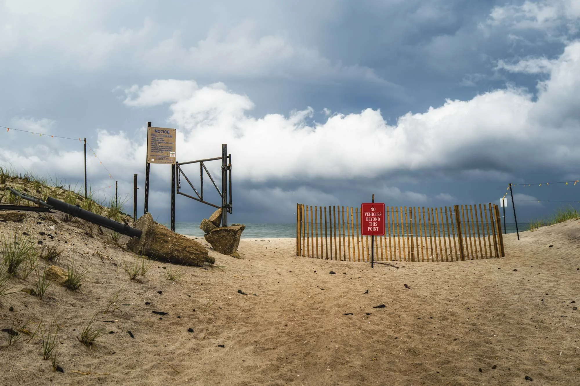 Beach entrance with warning signs, a sand dune, a wood fence blocking the path, and stormy clouds in the sky.