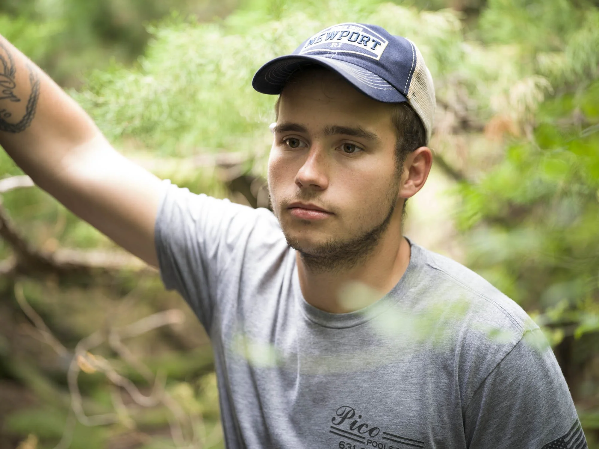 Young man wearing a blue and white baseball cap and a gray T-shirt with text, standing outdoors in a wooded area with green foliage.