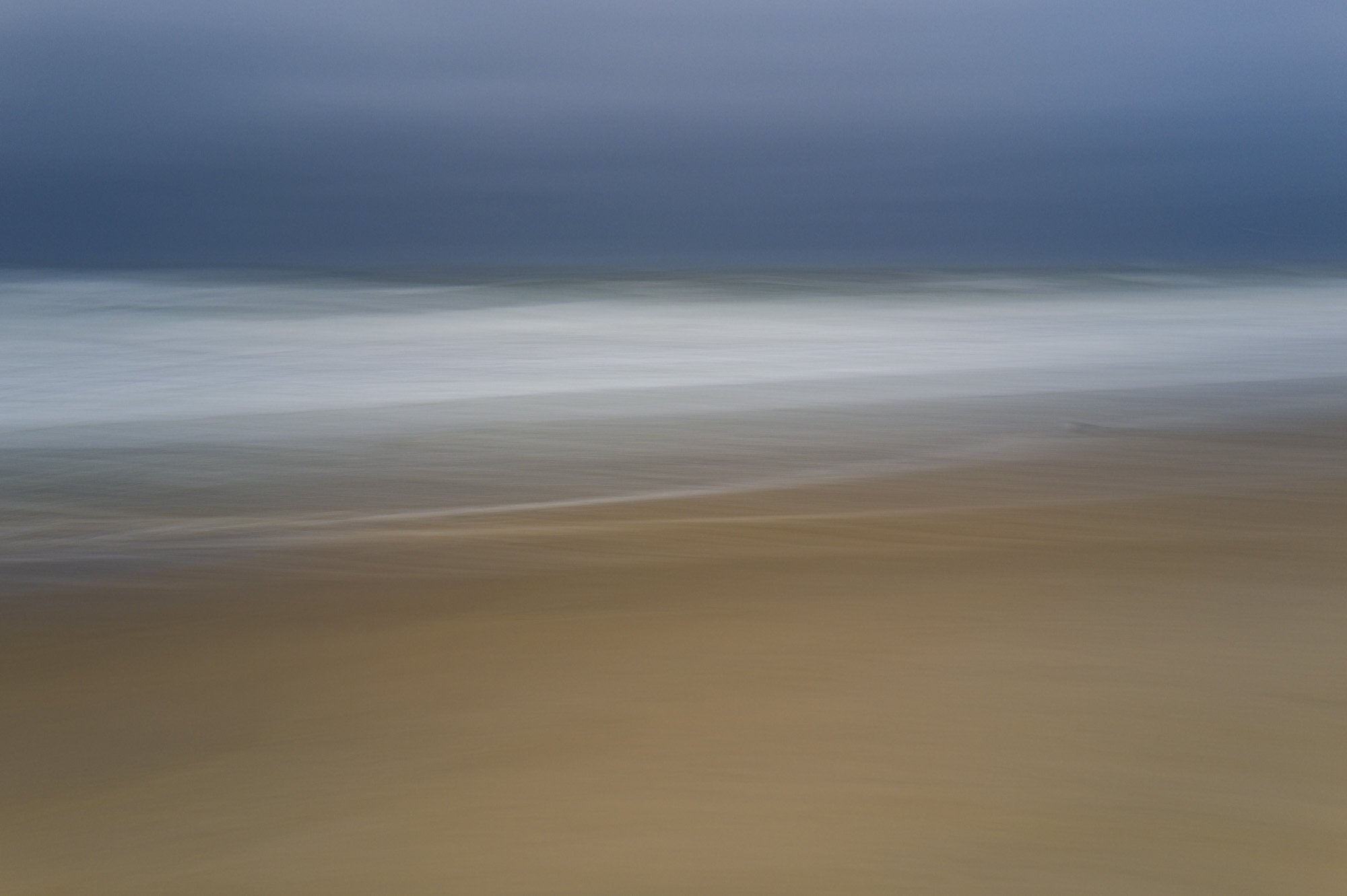 Blurred image of a beach with sand in the foreground, ocean waves in the middle, and dark stormy clouds in the sky above.