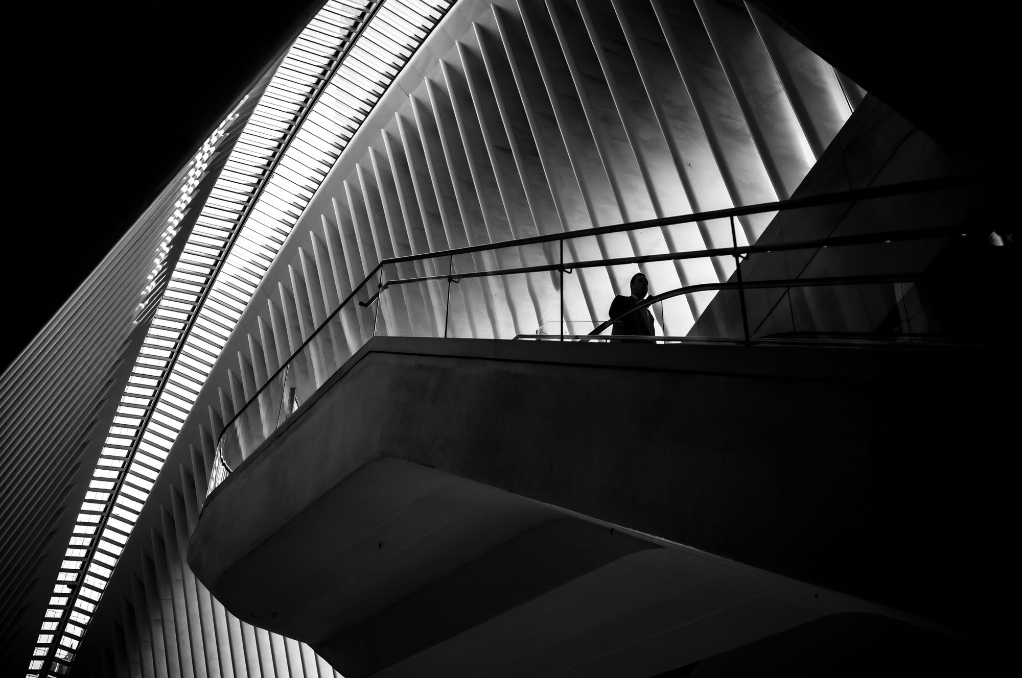 Black and white photo of a person standing on a balcony, with modern architectural building featuring curved lines and vertical panels in the background.