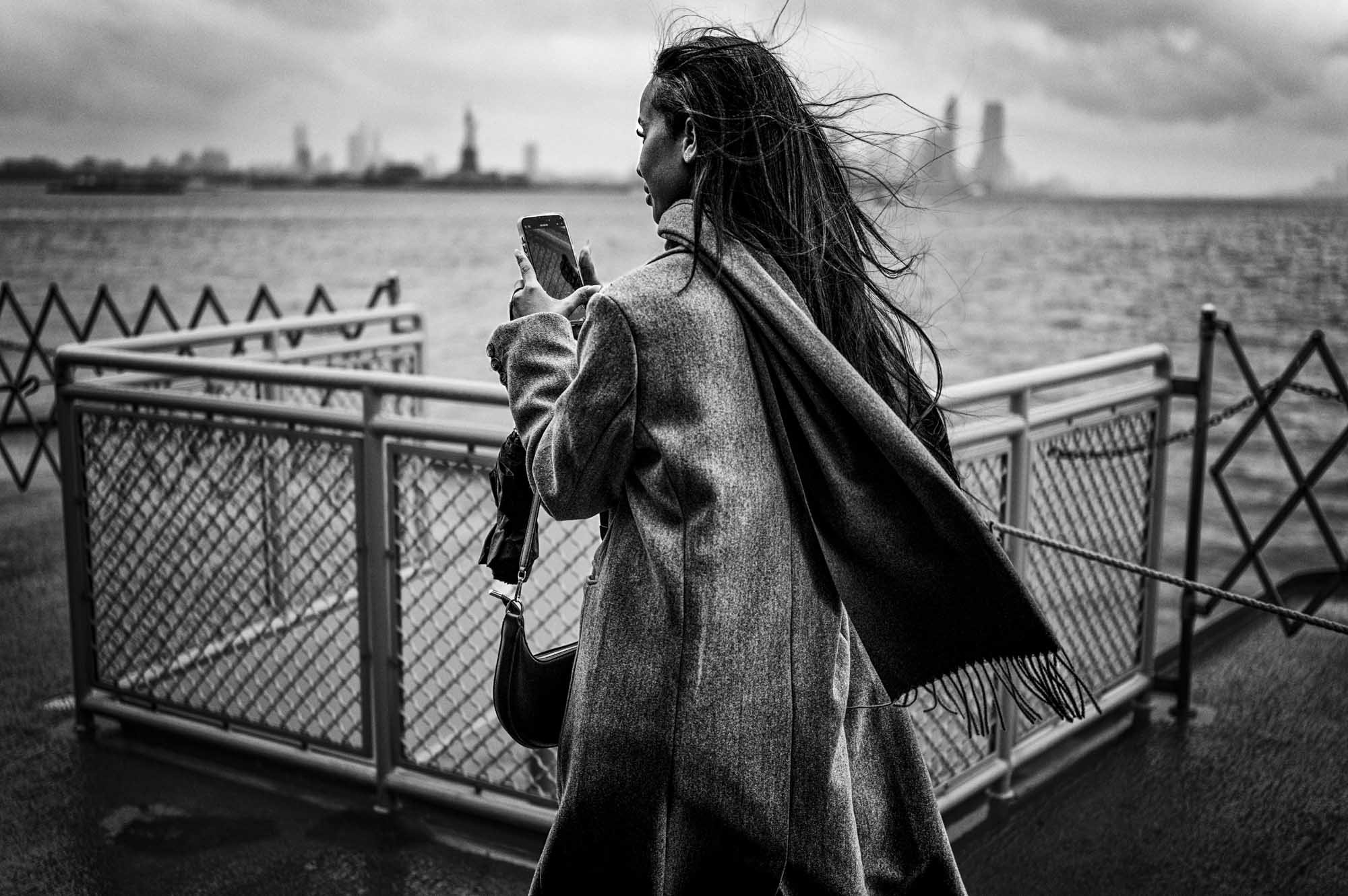 A woman wearing a coat, with long hair blown by the wind, standing near water, looking at her phone, with the Statue of Liberty and buildings in the background.