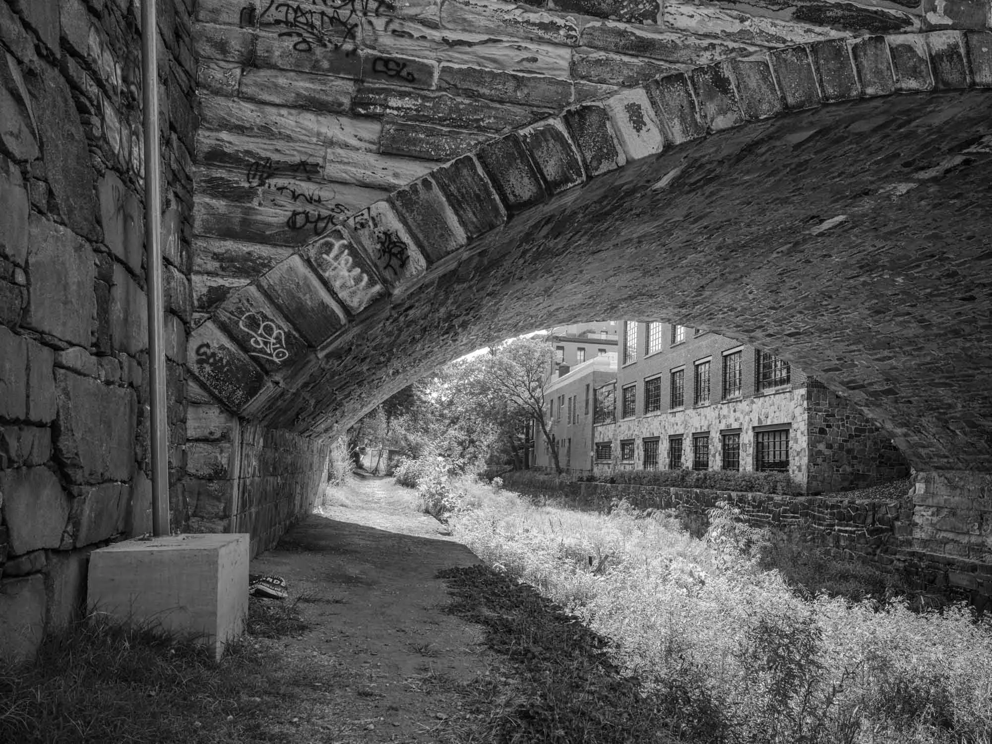 A black and white photo of a brick underpass with graffiti, a walking trail, and buildings in the background.