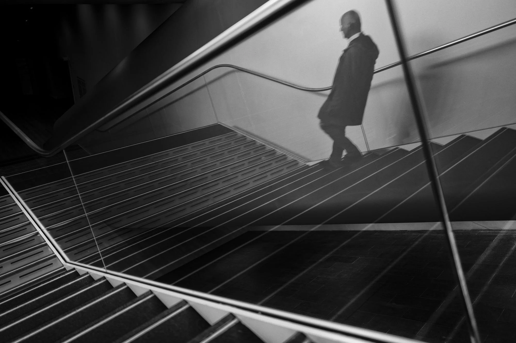 A black and white photo of a person walking down a stairway, with their reflection visible on a shiny, curved metallic surface beside them.