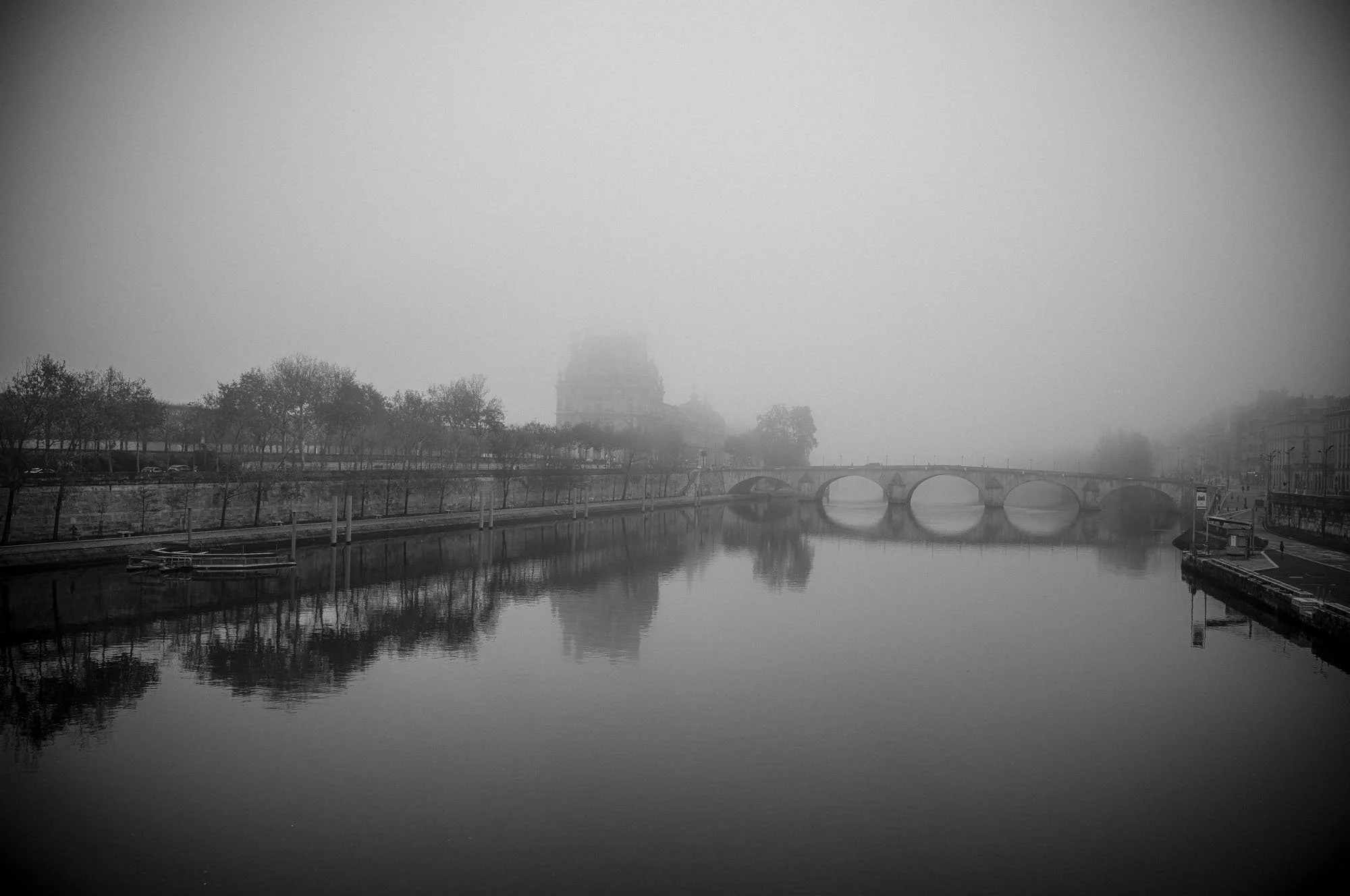 A black and white photo of a river with a stone bridge and a foggy cityscape in the background.