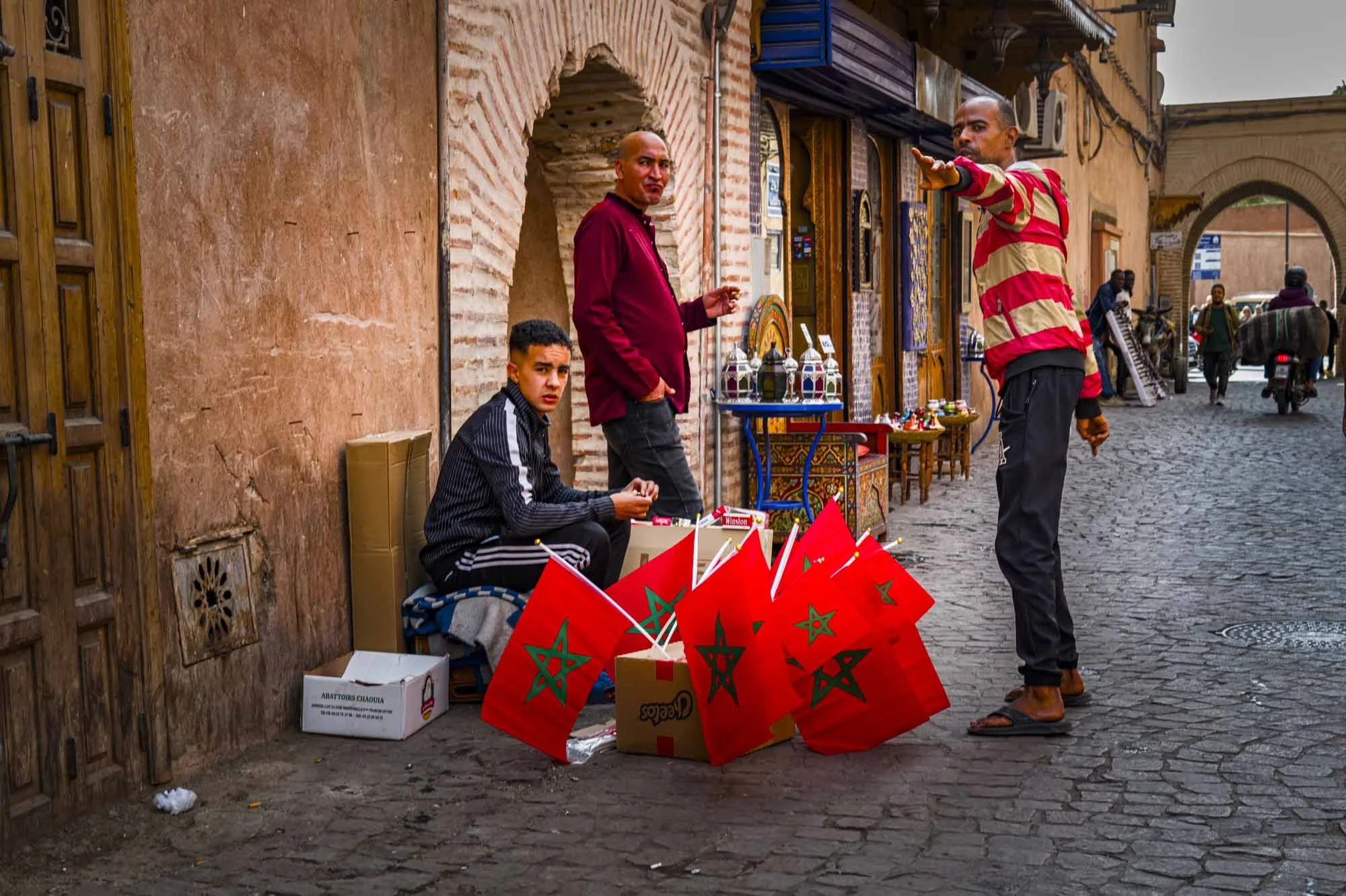 Street vendor selling Moroccan flags and souvenirs on a cobblestone street in a North African city, with three men engaging in conversation.