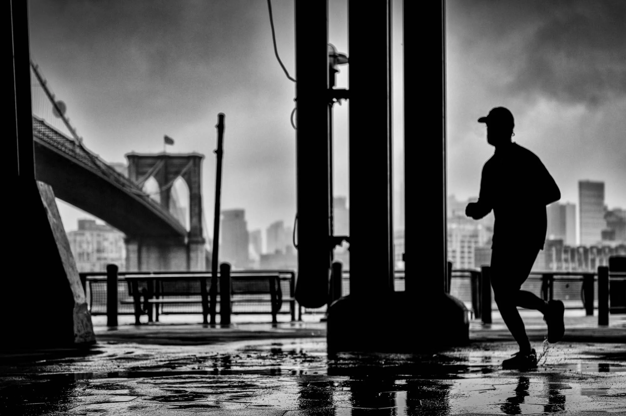 Silhouette of a man jogging on a wet pavement near the New York City skyline and Brooklyn Bridge during overcast weather.