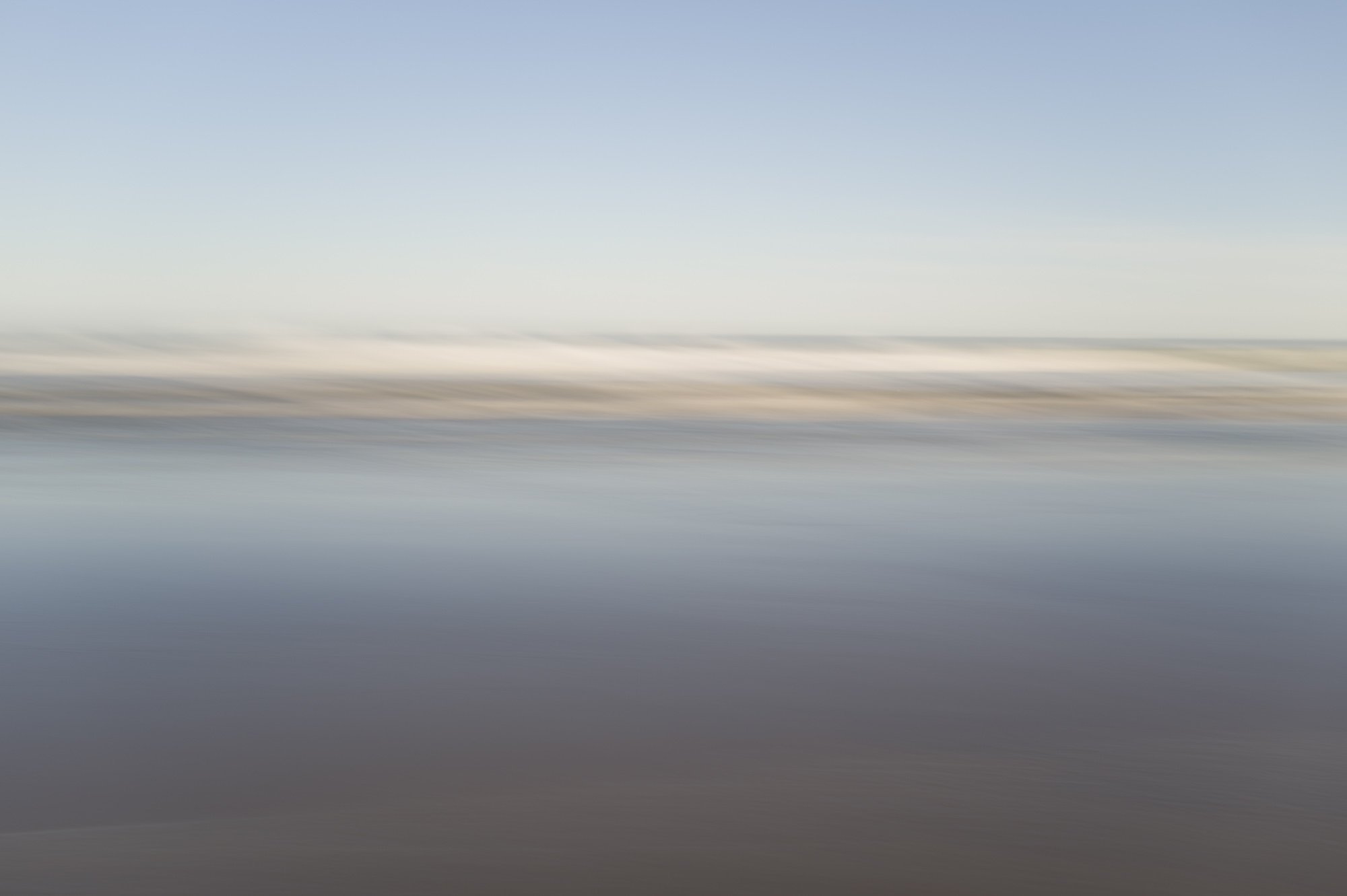 Blurred landscape of a body of water with a distant shoreline and a light blue sky.